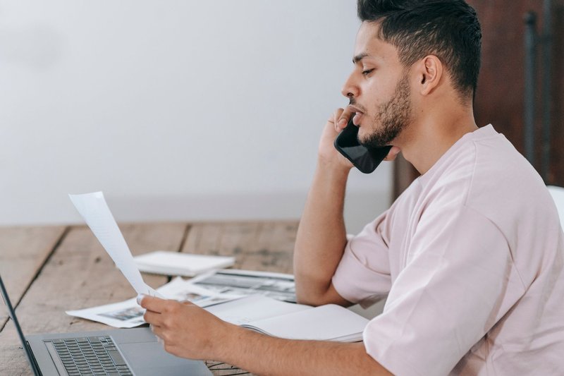 Junger Mann telefoniert am Holztisch, liest dabei ein Schreiben, daneben ein aufgeklappter Laptop und Unterlagen
