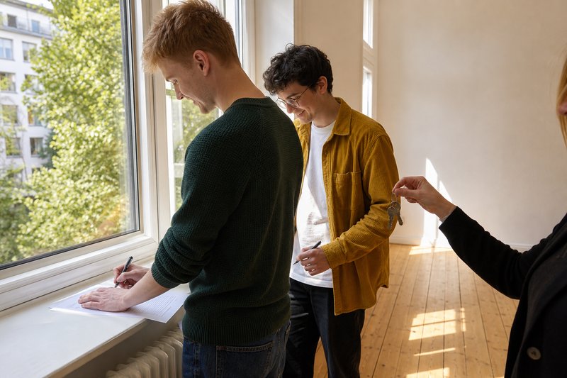 Zwei Studenten unterschreiben in einer leeren Altbauwohnung nacheinander denselben Mietvertrag, die Vermieterin hält die Schlüssel
