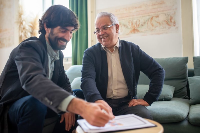 Ein Vertreter zeigt einem älteren Mann im Wohnzimmer auf dem Sofa ein Vertragsformular auf dem Beistelltisch