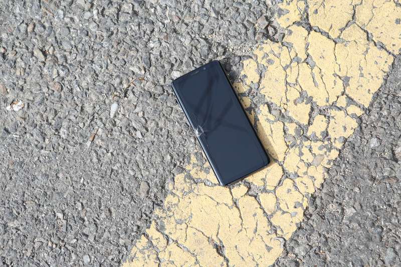 Smartphone mit gesprungenem Display liegt auf sonnigem Asphalt quer über einer gelben Fahrbahnmarkierung