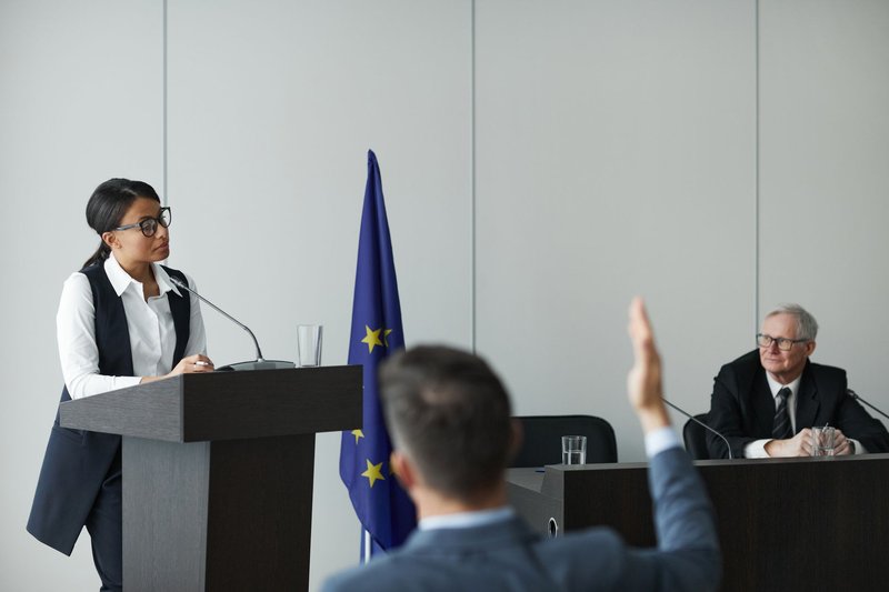 Rednerin steht an einem Pult neben einer Europaflagge, im Vordergrund hebt ein Zuhörer die Hand für eine Frage