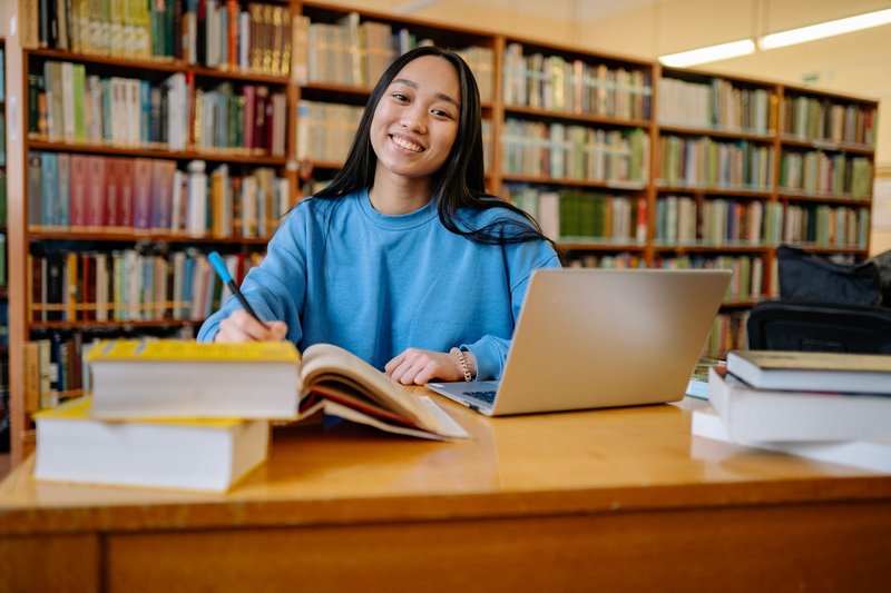 Studentin arbeitet am Laptop an einem Bibliothekstisch vor hohen Bücherregalen