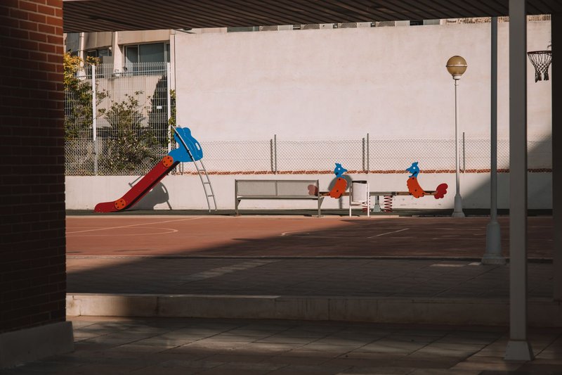 Leerer Spielplatz mit roter Rutsche und Federwippen auf gepflastertem Boden vor einer hellen Wand