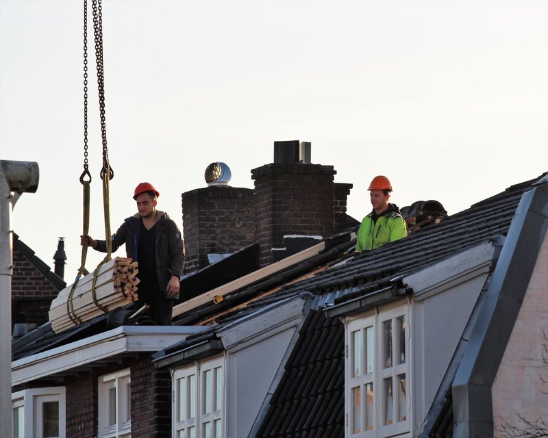 Zwei Bauarbeiter mit Helm nehmen auf einem abgedeckten Hausdach ein am Kranhaken hängendes Lattenbündel an