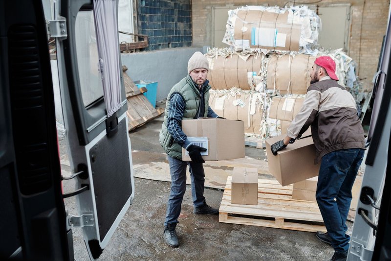 Zwei Arbeiter heben Kartons in einen Lieferwagen, daneben steht eine Palette auf einem Hof
