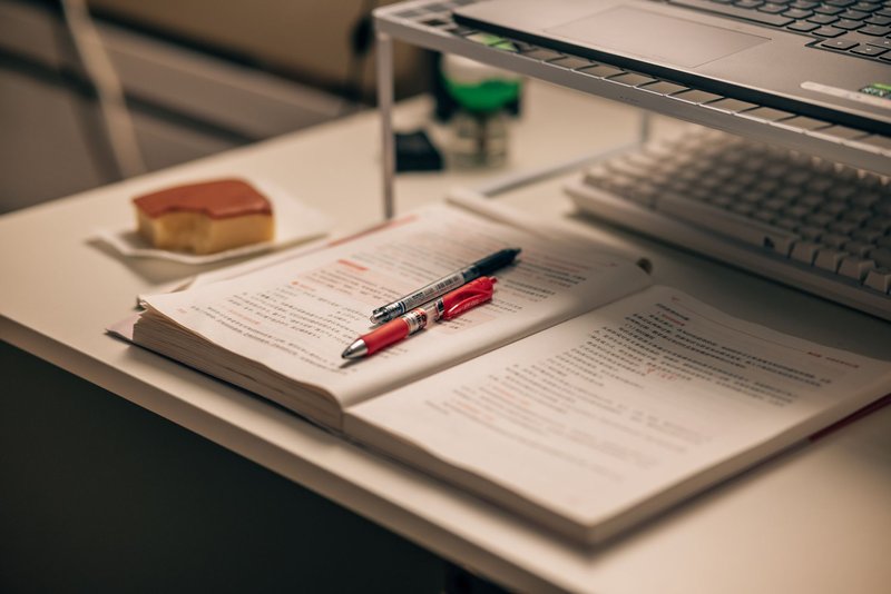 Aufgeschlagenes, mit rotem Stift markiertes Buch auf einem Schreibtisch, daneben ein Laptop