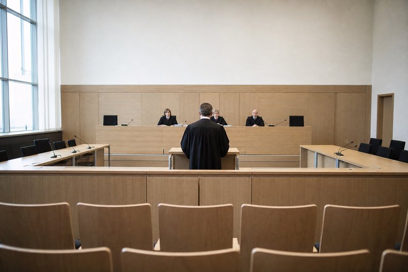 Blick aus dem Zuschauerraum auf die Richterbank eines deutschen Gerichtssaals, davor ein Anwalt in Robe am Pult