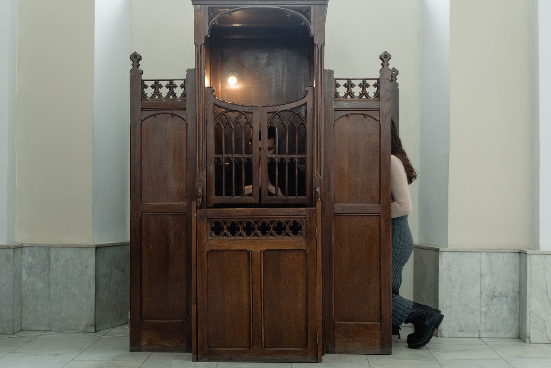 Hölzerner Beichtstuhl in einer Kirche, seitlich davor kniet eine Person mit abgewandtem Gesicht im Gespräch