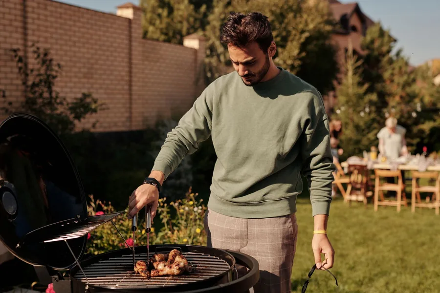 Mann grillt abends im Garten, im Hintergrund sitzen mehrere Personen an einem Tisch