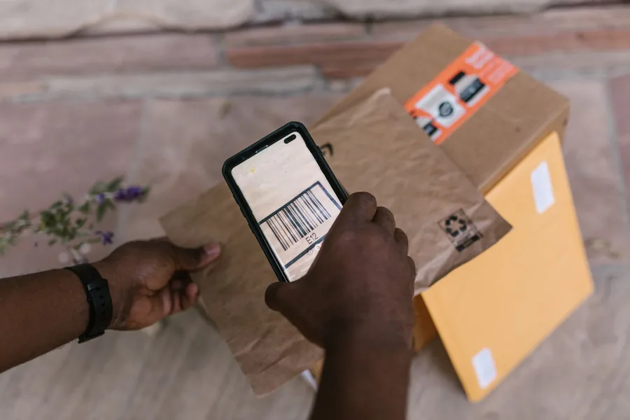 Hände scannen mit einem Smartphone den Barcode eines Pakets vor einer Haustür
