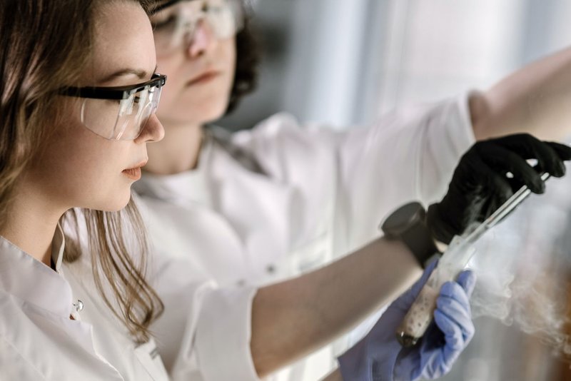Zwei Wissenschaftlerinnen mit Schutzbrillen führen im Labor einen Versuch mit einem Reagenzglas durch