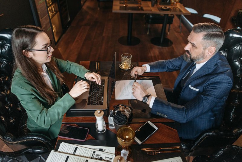 Ein Mann im Anzug und eine Frau im grünen Blazer besprechen bei einem Essen im Restaurant Unterlagen am Tisch