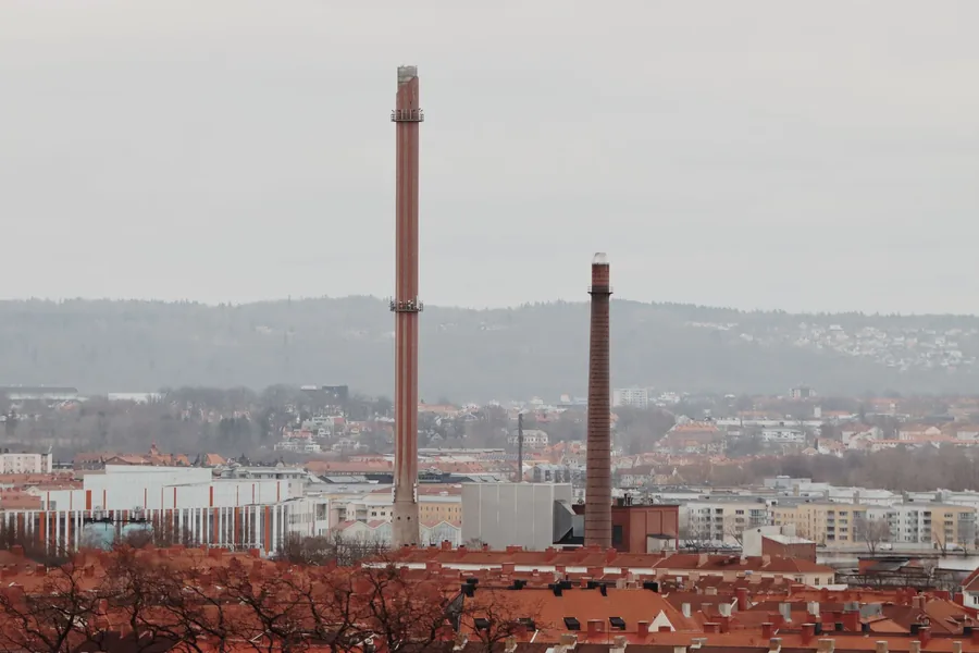 Zwei hohe Industrieschornsteine ragen direkt hinter dicht stehenden Wohnhäusern mit roten Ziegeldächern auf.