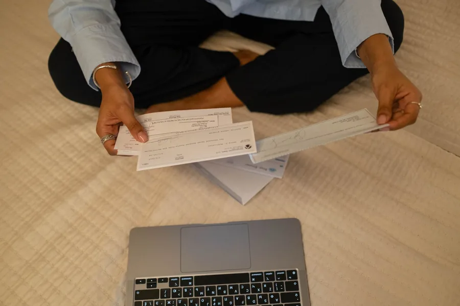 Person hält Schecks in den Händen neben einem aufgeklappten Laptop.