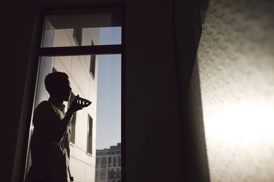 Die Silhouette eines Mannes am Fenster, der abends mit einem Smartphone am Ohr telefoniert