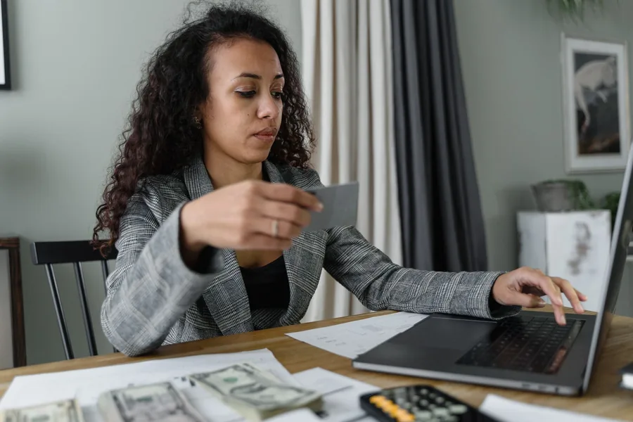 Junge Frau sitzt mit Laptop, Bargeld und Karte an einem Tisch und erledigt eine Überweisung.