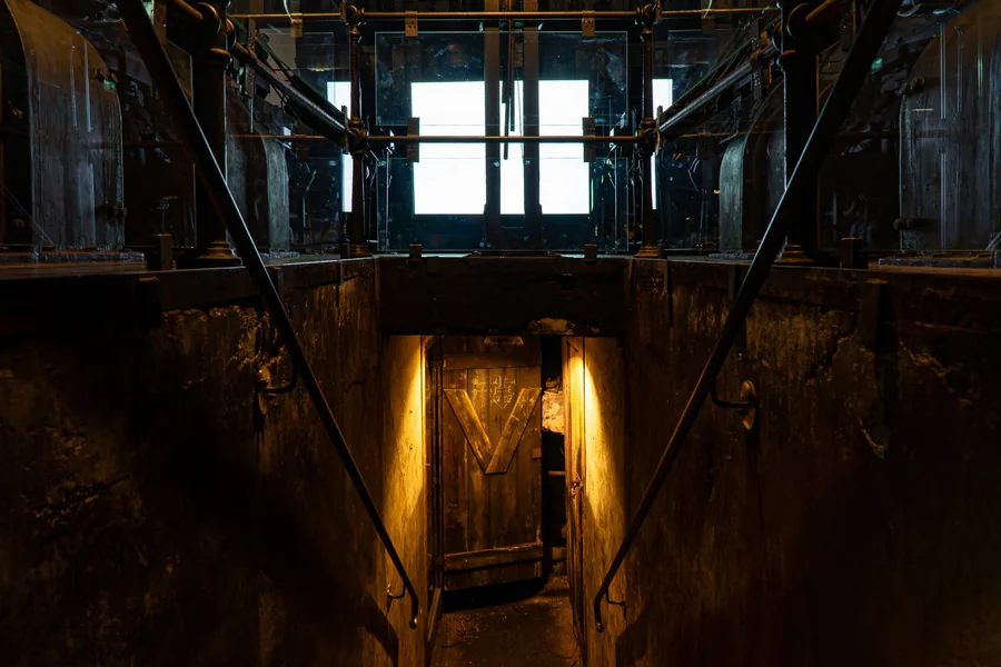 Blick von oben in ein dunkles Treppenhaus, unten fällt Licht durch eine offene Tür