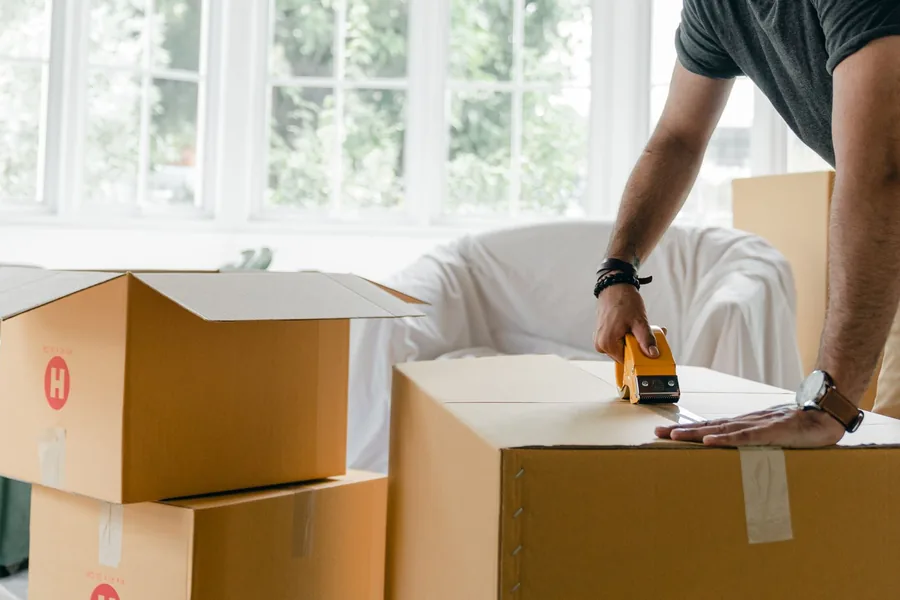 Eine Person verschließt einen Umzugskarton mit Klebeband in einem hellen Zimmer.