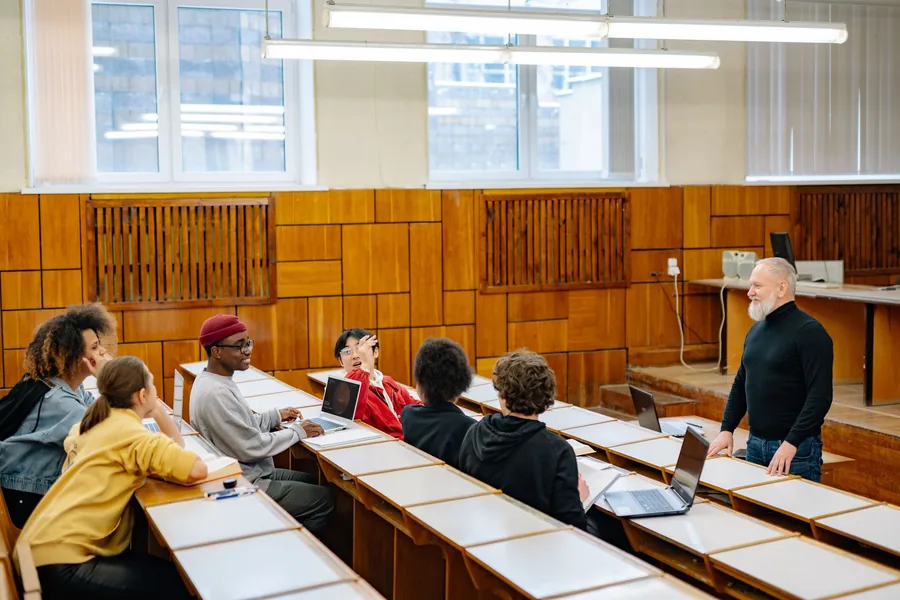Dozent steht vor Studierenden in einem holzvertäfelten Seminarraum und bespricht mit ihnen ihre Arbeiten