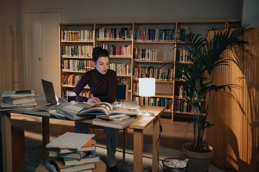 Eine Studentin lernt abends allein am Schreibtisch, umgeben von aufgeschlagenen Büchern, mit einer warmen Schreibtischlampe und einem Bücherregal im Hintergrund
