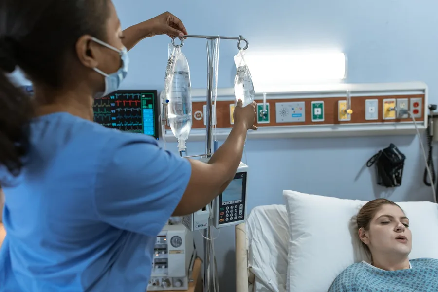 Pflegekraft in Schutzkleidung stellt am Krankenbett eine Infusionspumpe ein, im Hintergrund liegt eine Patientin