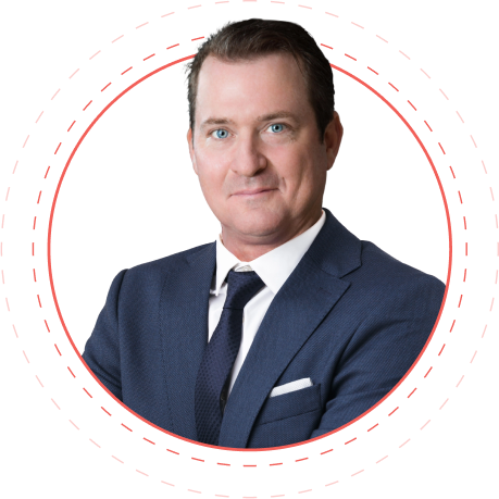 A man wearing a black suit and tie with a white shirt poses for a professional headshot. There are circles framing him. 