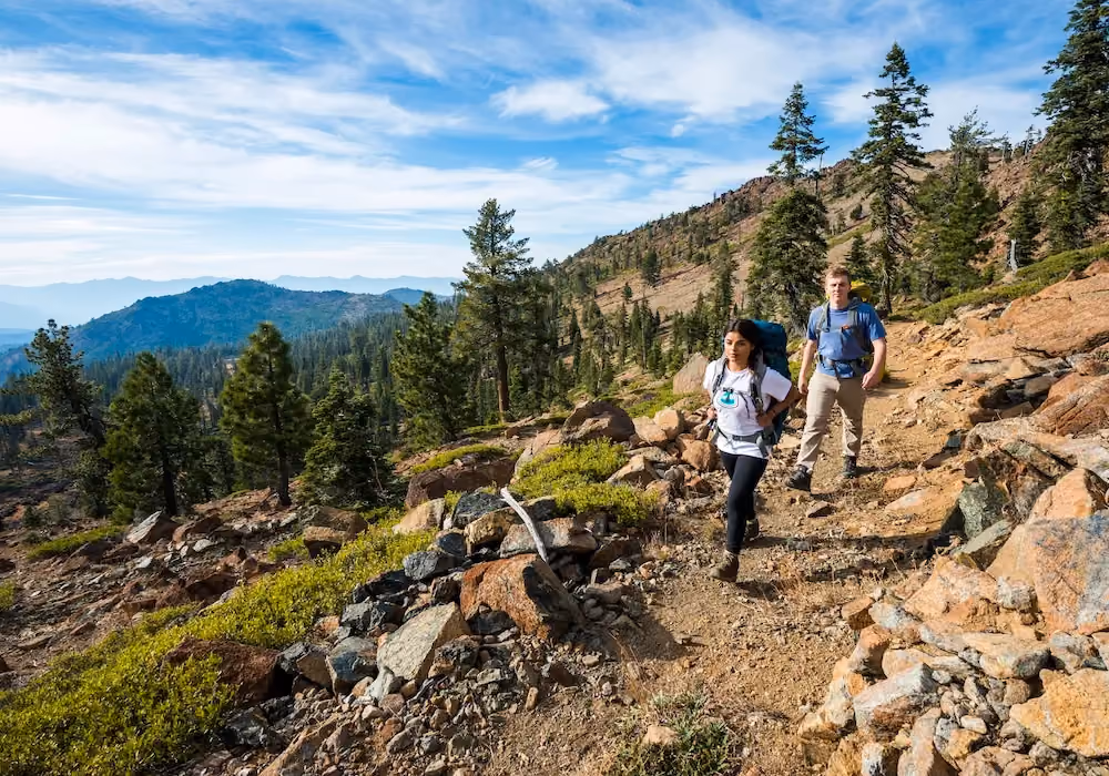 Two hikers with backpacks walking on a rocky mountain trail surrounded by pine trees under a blue sky.