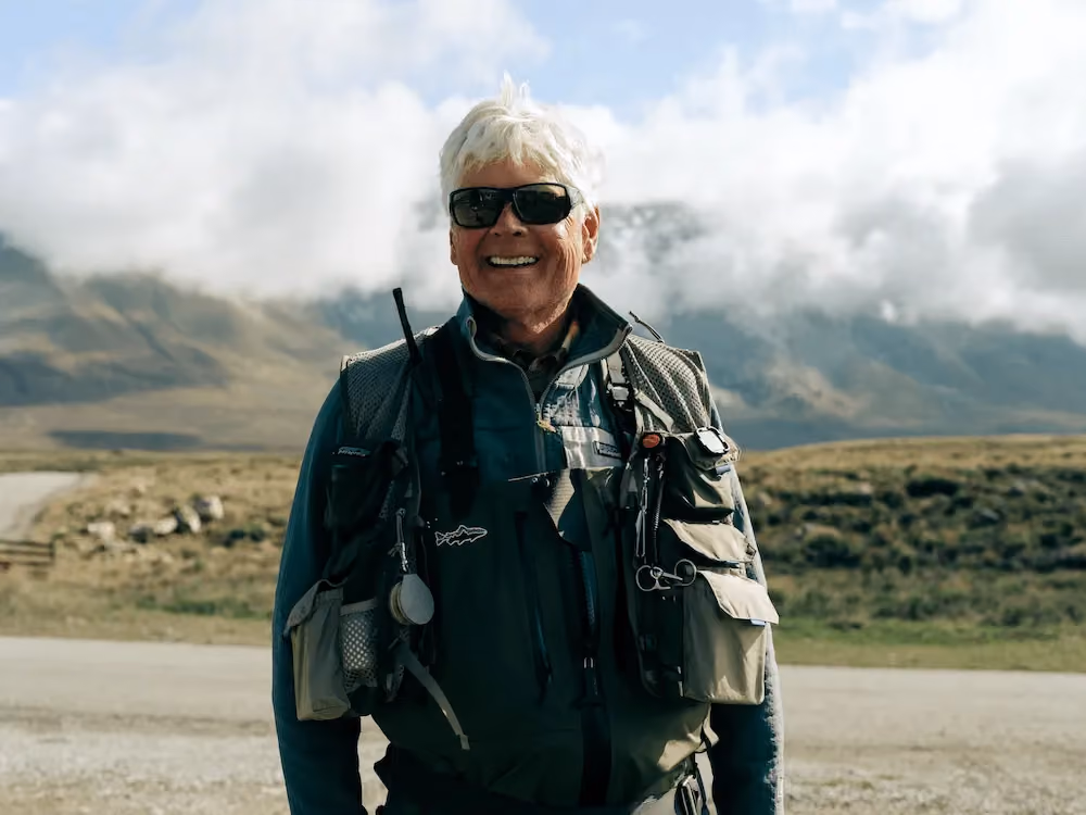 Smiling older man wearing sunglasses and fishing gear outdoors with mountains and clouds in the background.