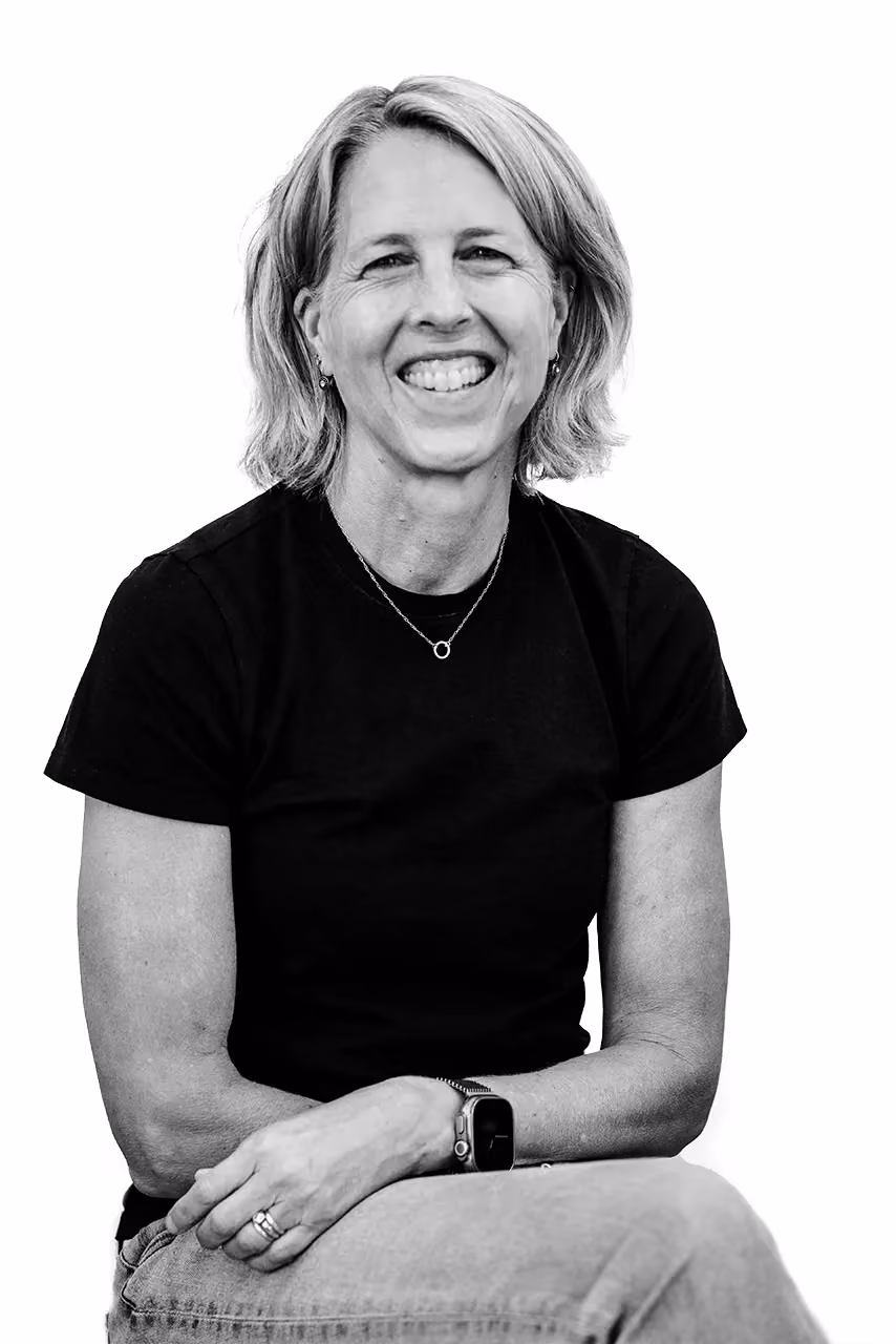 Middle-aged woman with short hair smiling, wearing a black shirt and jeans, sitting with arms crossed.