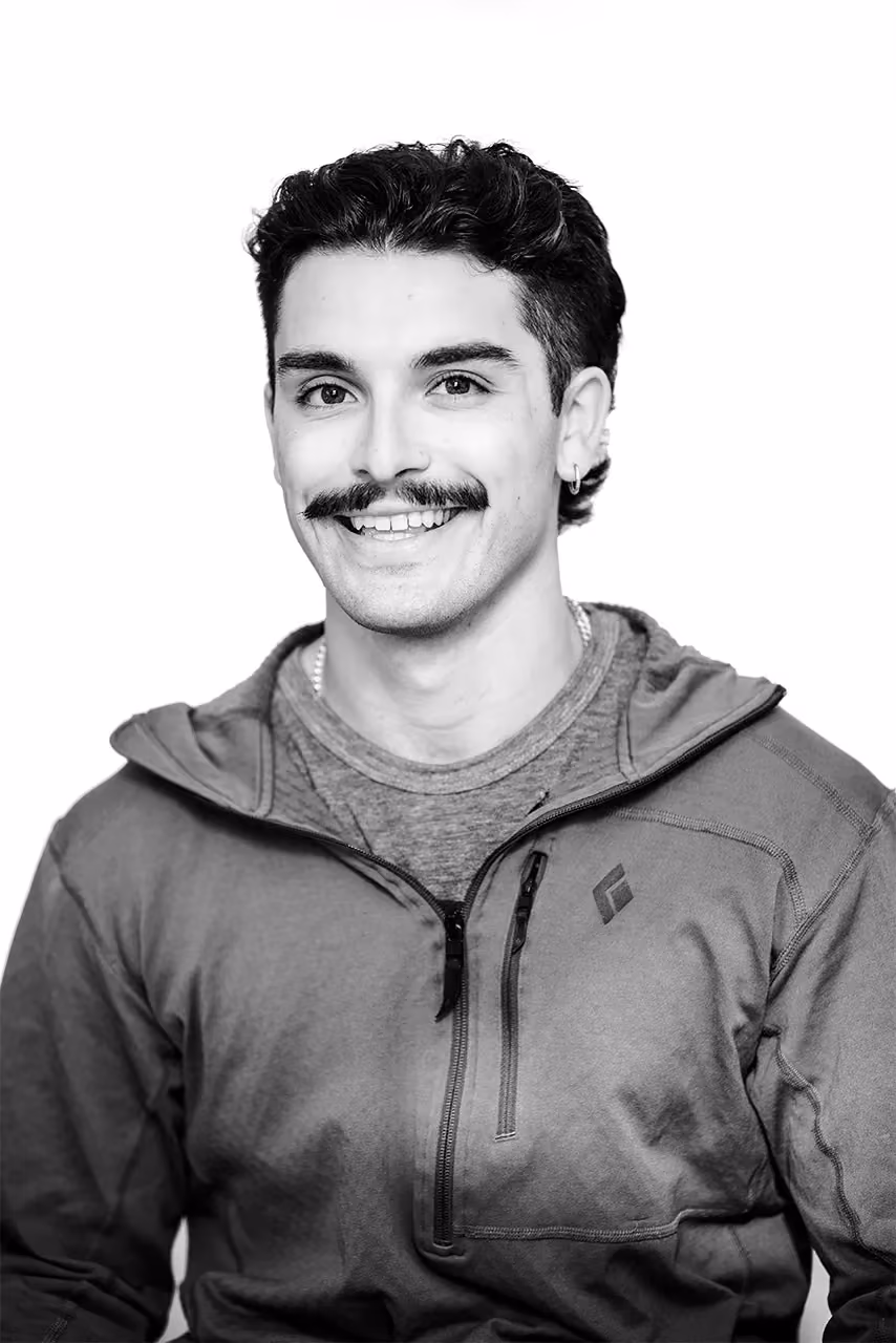 Portrait of a smiling man with dark curly hair, mustache, and wearing a zip-up jacket and earring.