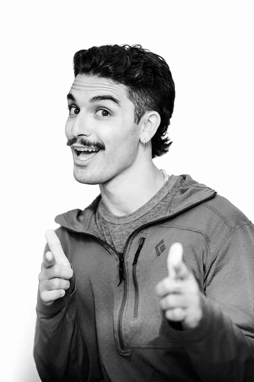 Man with mustache and hoop earring pointing finger guns and smiling in a jacket with zipper.