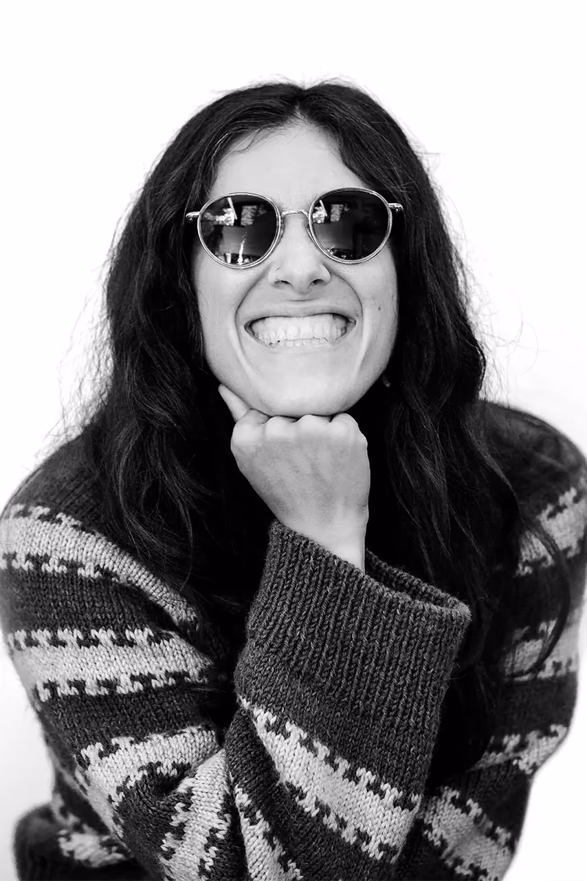 Smiling person wearing round sunglasses and a patterned sweater resting their chin on their fist.