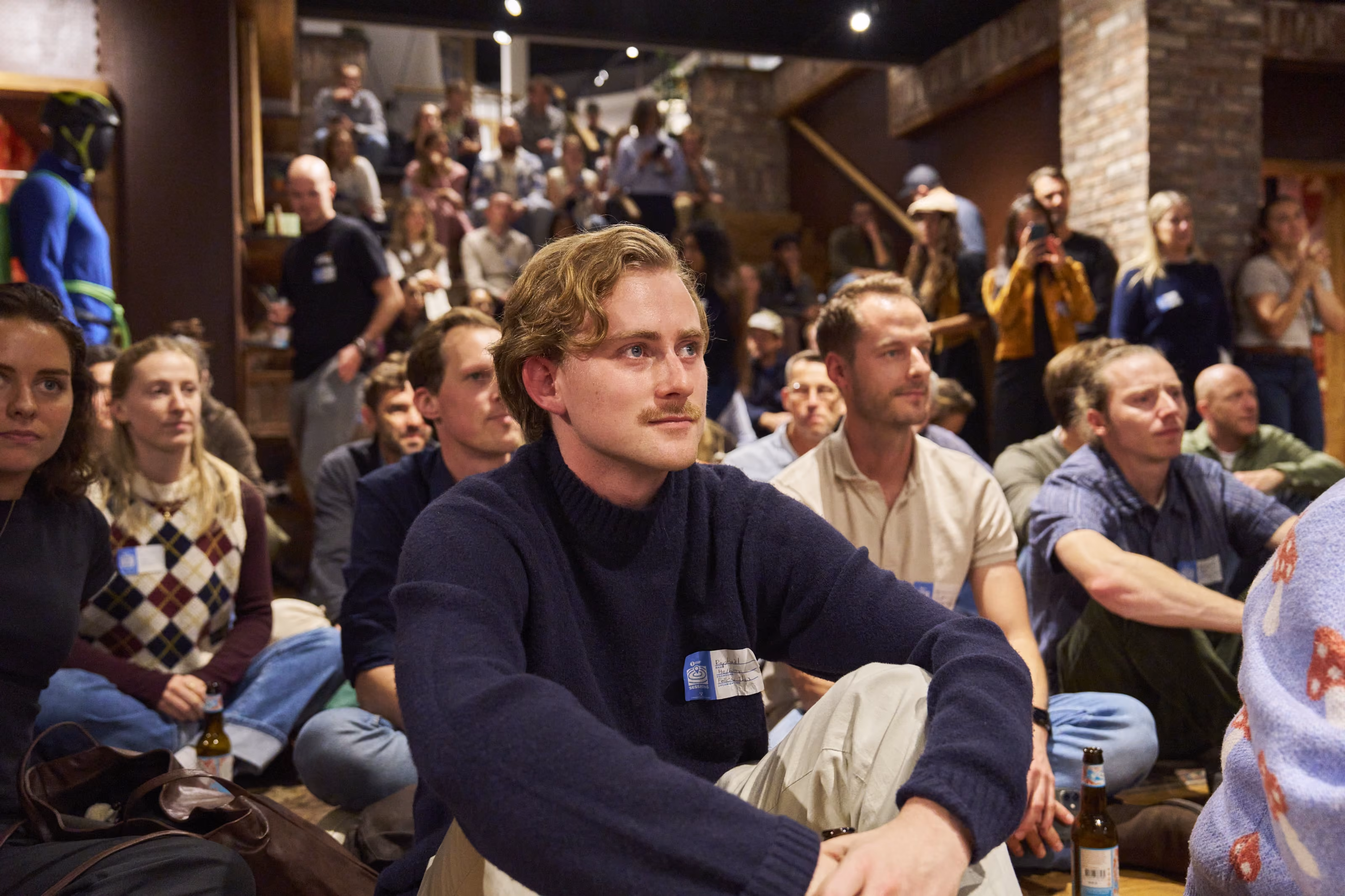 Group of people sitting on floor and stairs attentively listening to an event in a rustic room.