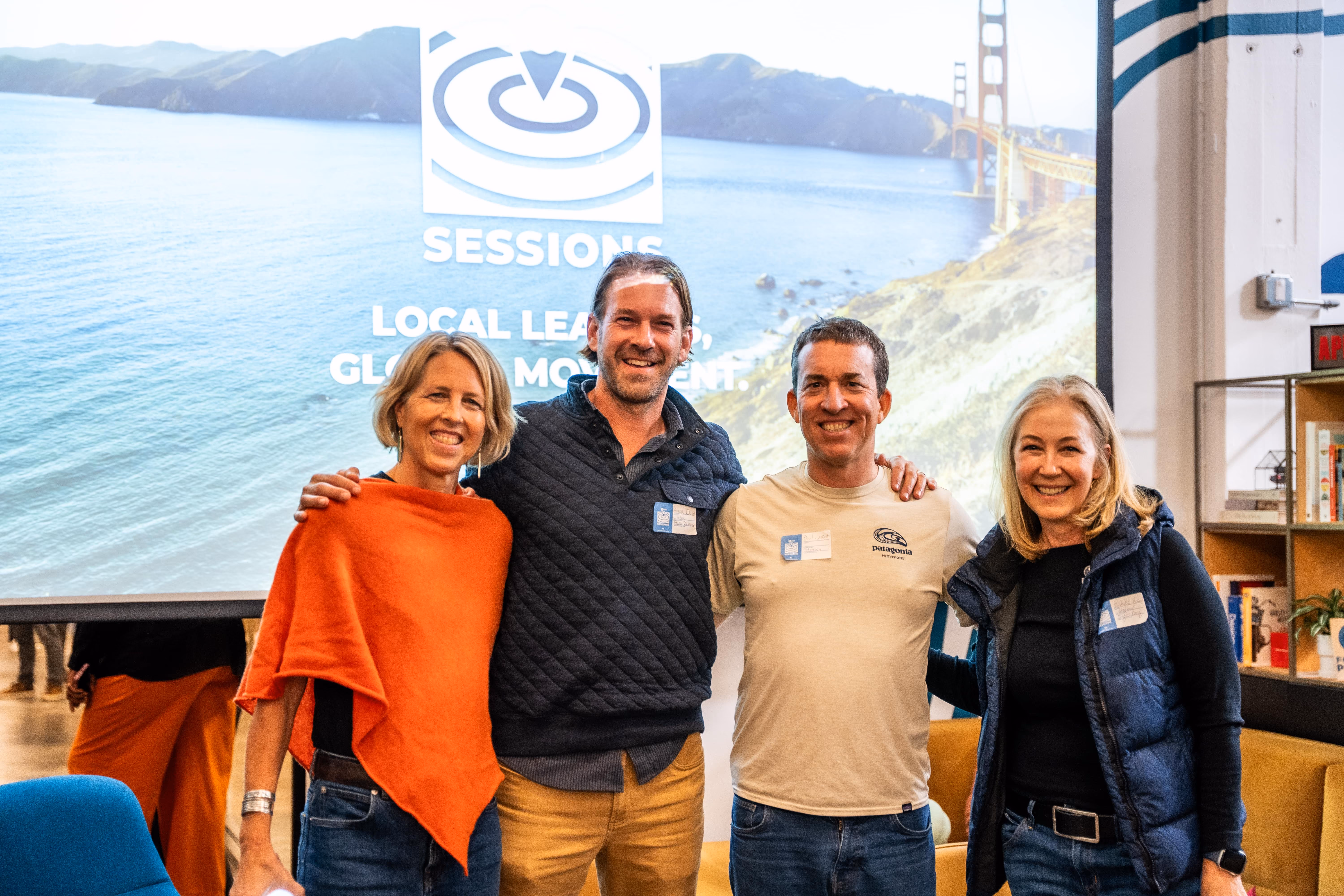 Four smiling adults posing with arms around each other in front of a screen with ocean and bridge image.