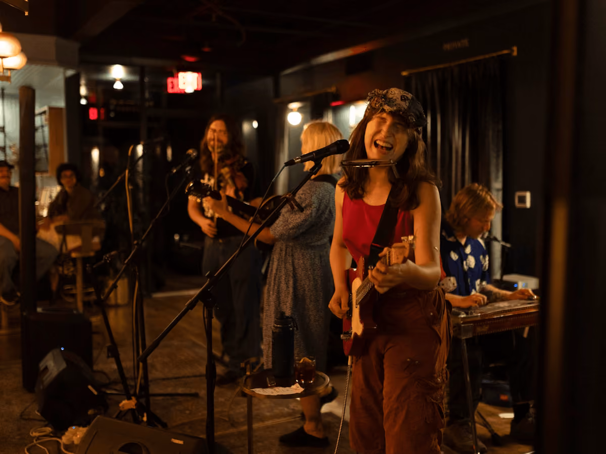 Band playing live indoors: woman in red sings with guitar and harmonica, others play violin, steel guitar.
