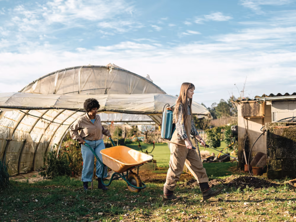 Two women work outdoors near a greenhouse; one pushes a wheelbarrow, the other sprays plants with a backpack sprayer.