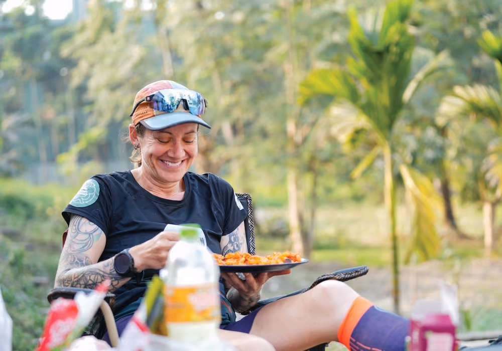 Smiling woman with tattoos sitting outdoors holding a plate of food with trees in background.