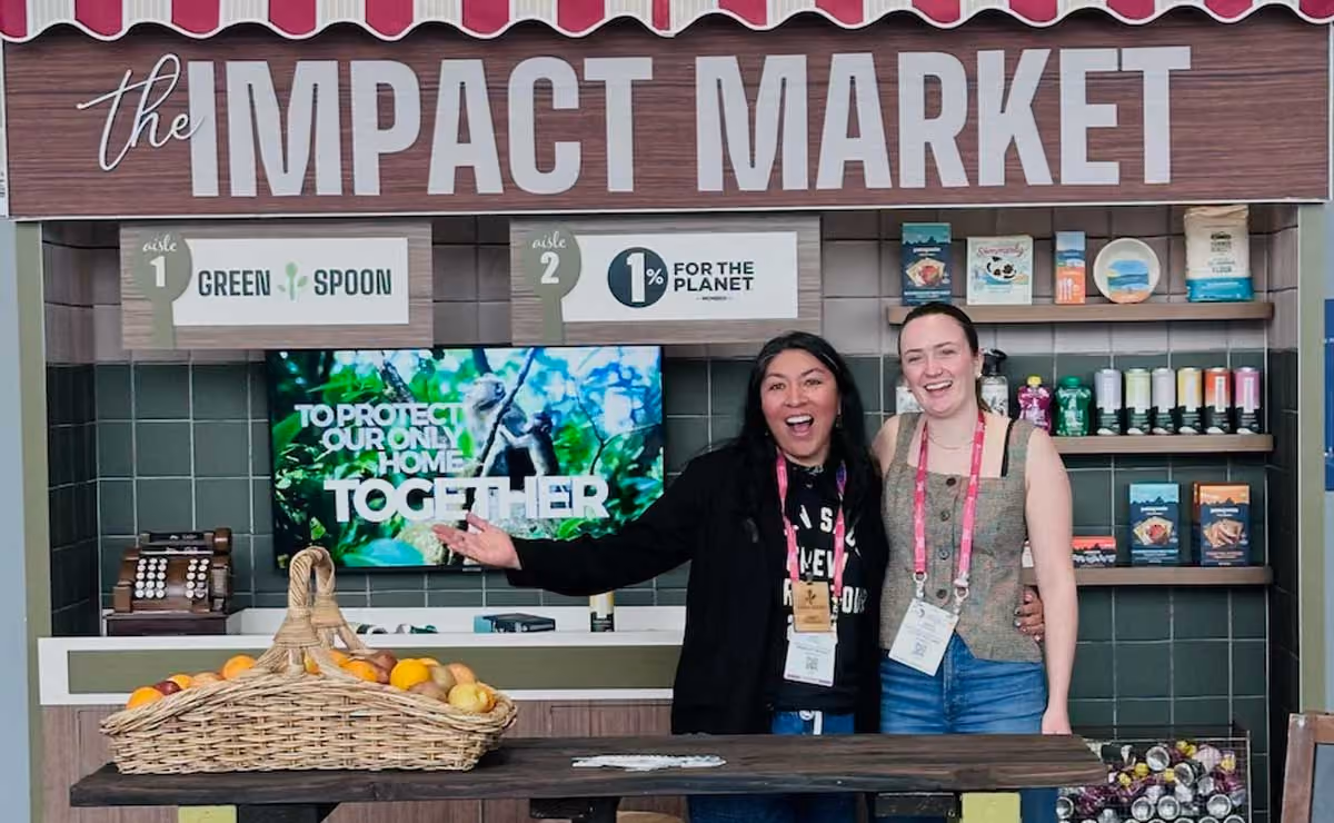 Two smiling women standing behind a market stall with baskets of fruit under a sign reading 'the IMPACT MARKET' with product shelves and a digital display in the background.
