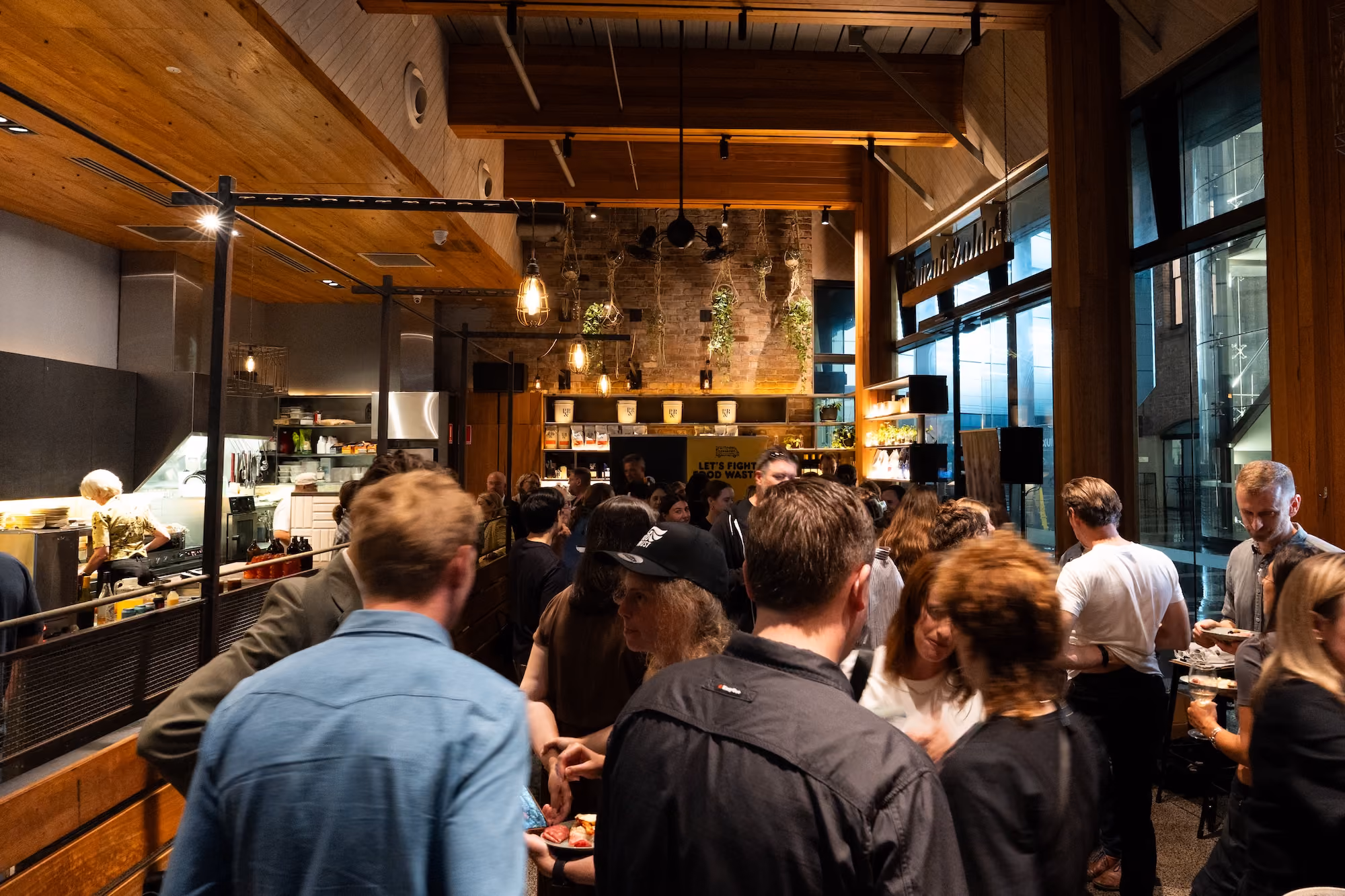 Crowded indoor gathering at a restaurant or cafe with warm wooden decor, exposed brick wall, hanging plants, and pendant lights, with people socializing and some holding plates of food and drinks.