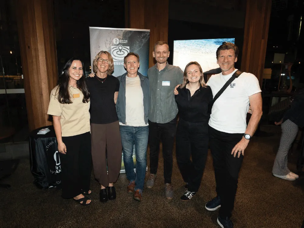 Group of six people standing indoors smiling in front of a banner and a screen at an event.