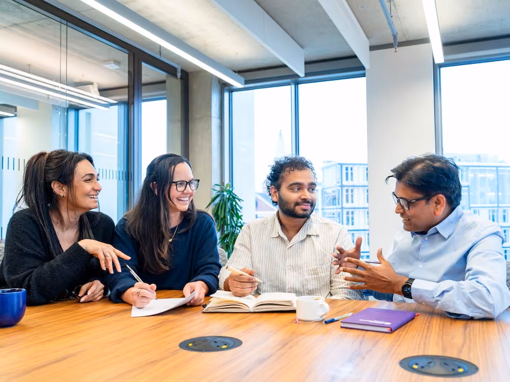 Four diverse colleagues sitting at a table with notebooks, discussing in a bright office space.