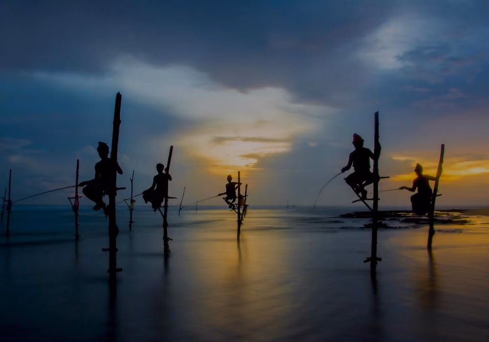 Silhouettes of five fishermen sitting on stilts in the water, fishing at sunset under a cloudy sky.