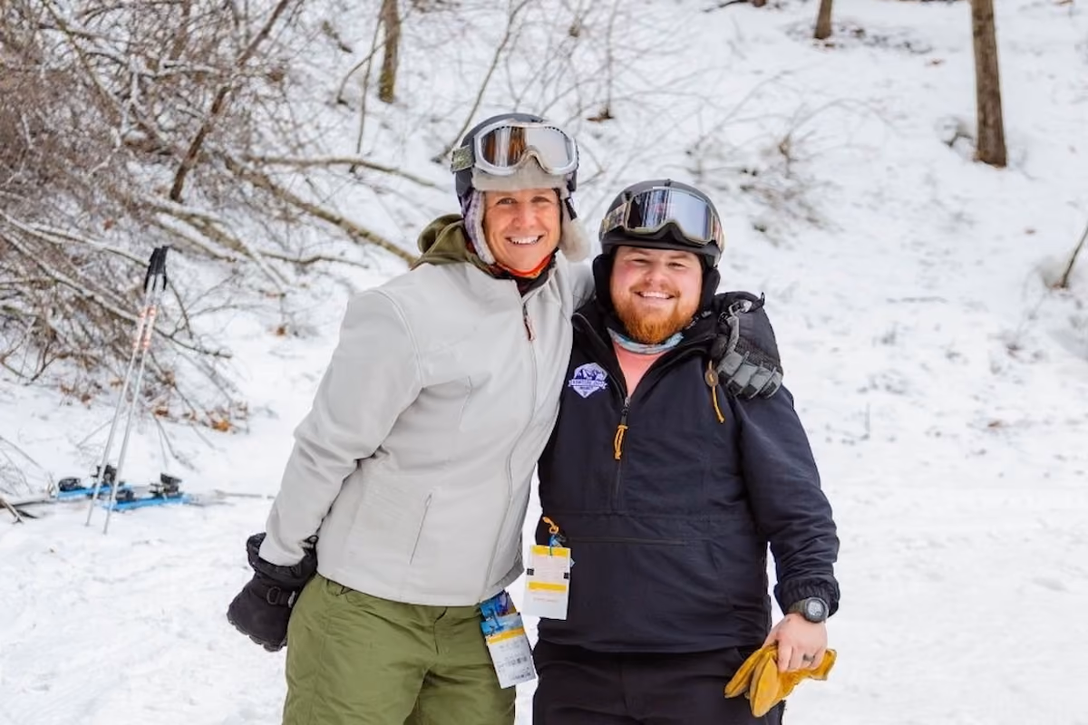 Two people in winter ski gear standing close together and smiling in a snowy outdoor setting, with ski poles and skis resting against a tree in the background.