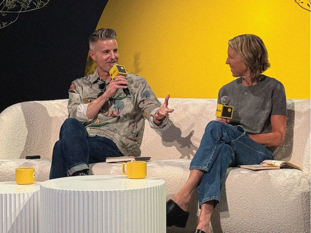 Two people seated on a white couch, holding yellow microphones, talking in a studio with yellow background.