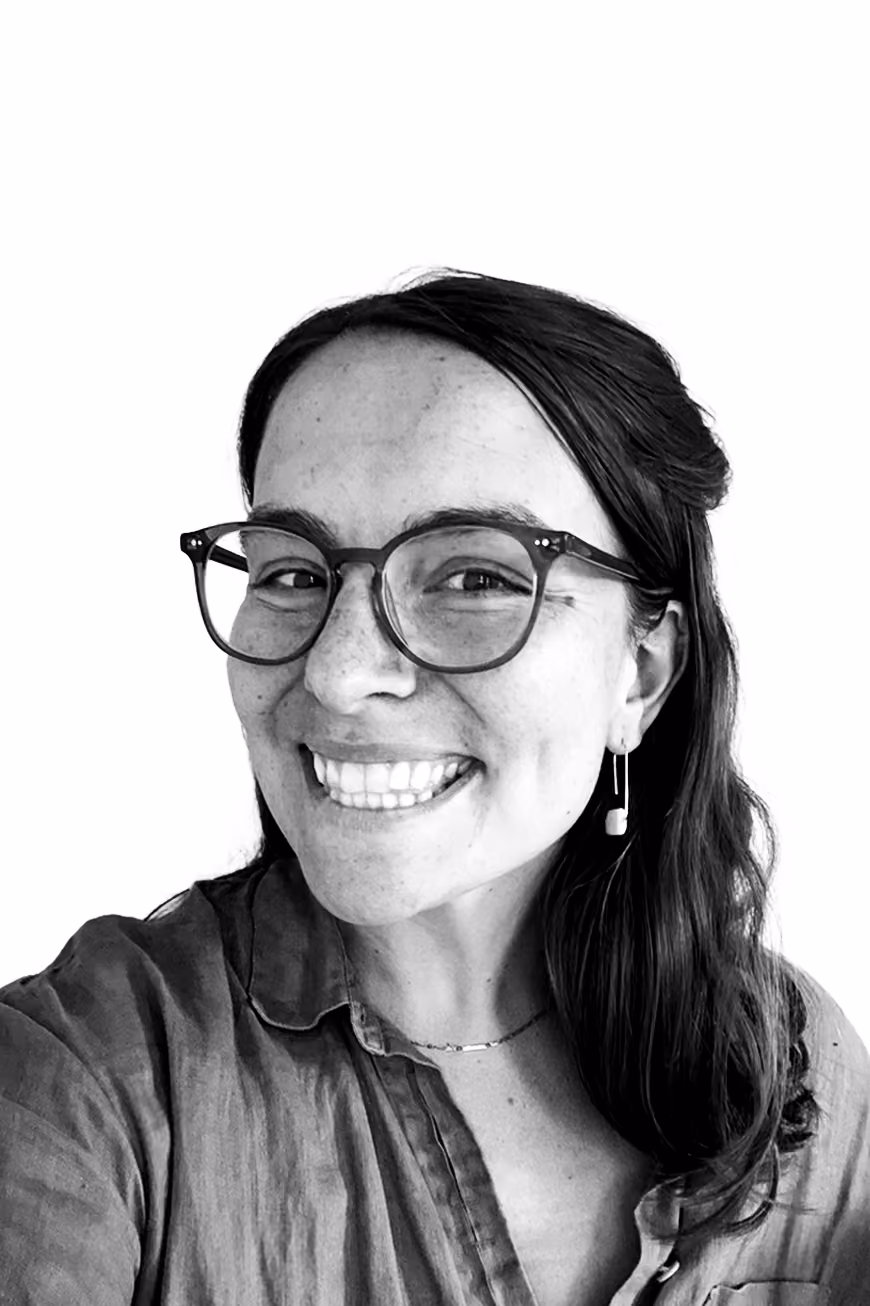 Smiling woman with glasses, long dark hair, wearing earrings and a collared shirt, against a white background.