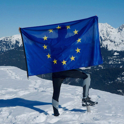 Person standing on a snowy mountain, holding the EU flag.