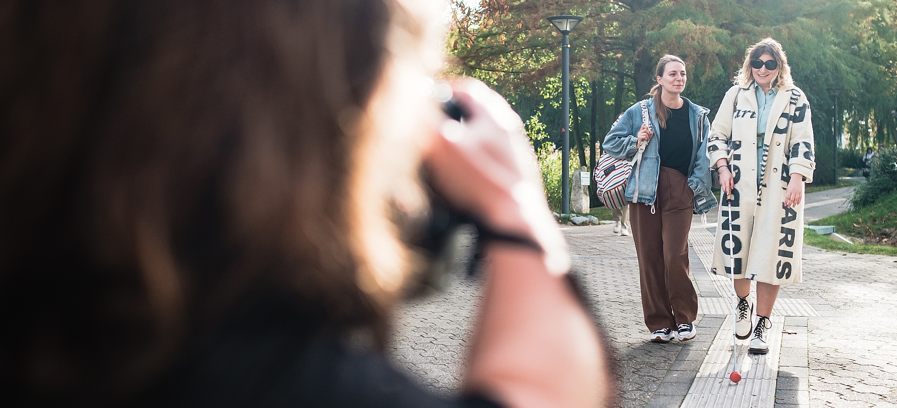 Zwei Frauen spazieren in der Sonne durch einen Park und werden fotografiert. Eine von ihnen orientiert sich mit einem Langstock am Bodenleitstreifen zum Tasten. Das Foto nimmt die Perspektive der Fotografin ein. Man sieht den Rücken der Fotografin: Sie ist kleinwüchsig.