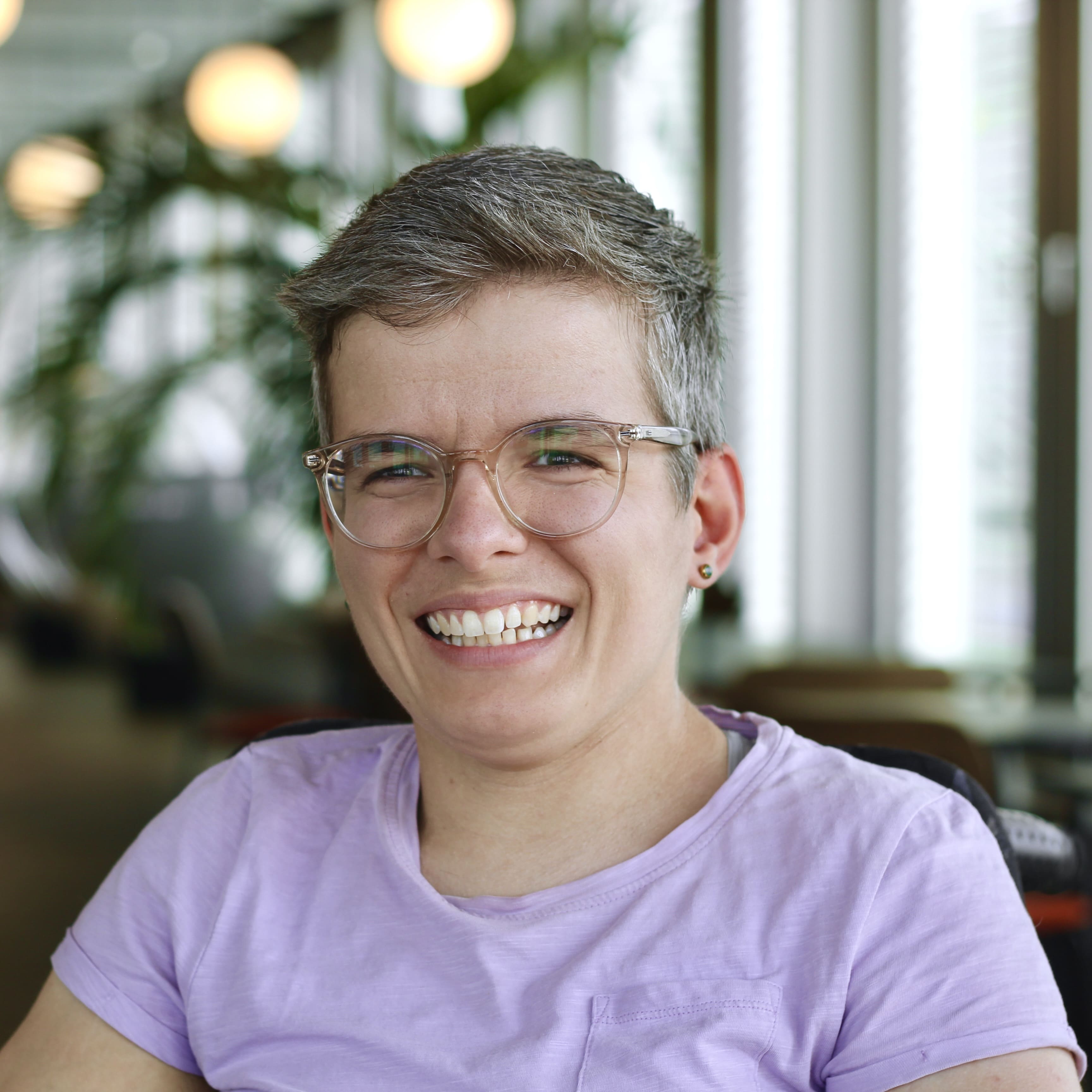 Viktoria hat kurze graumelierte Haare, trägt eine Brille und ein fliederfarbenes T-Shirt