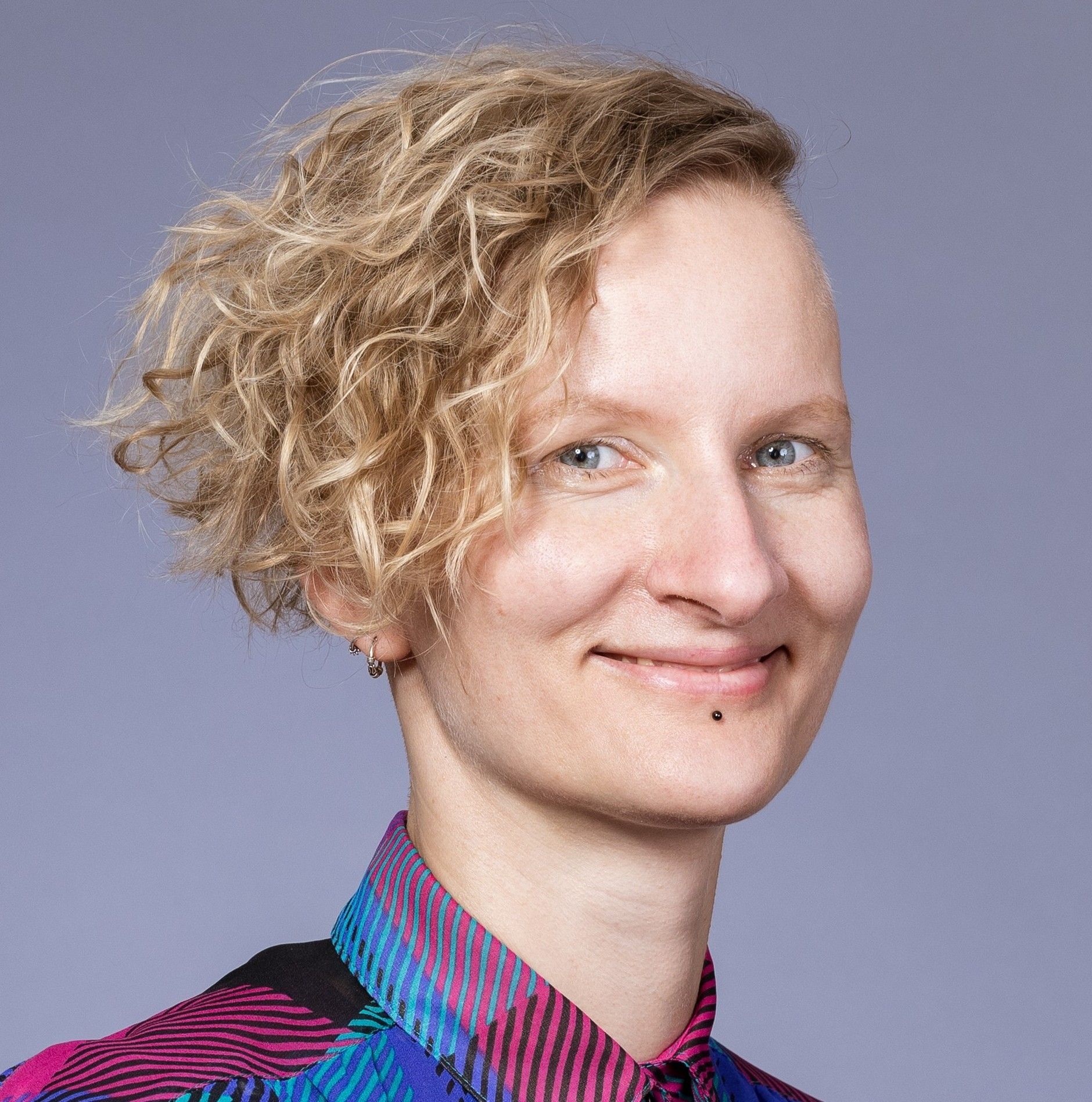 Porträtfoto von Luise. Sie hat mittellange lockige blonde Haare, trägt ein karriertes Oberteil und lächelt in die Kamera