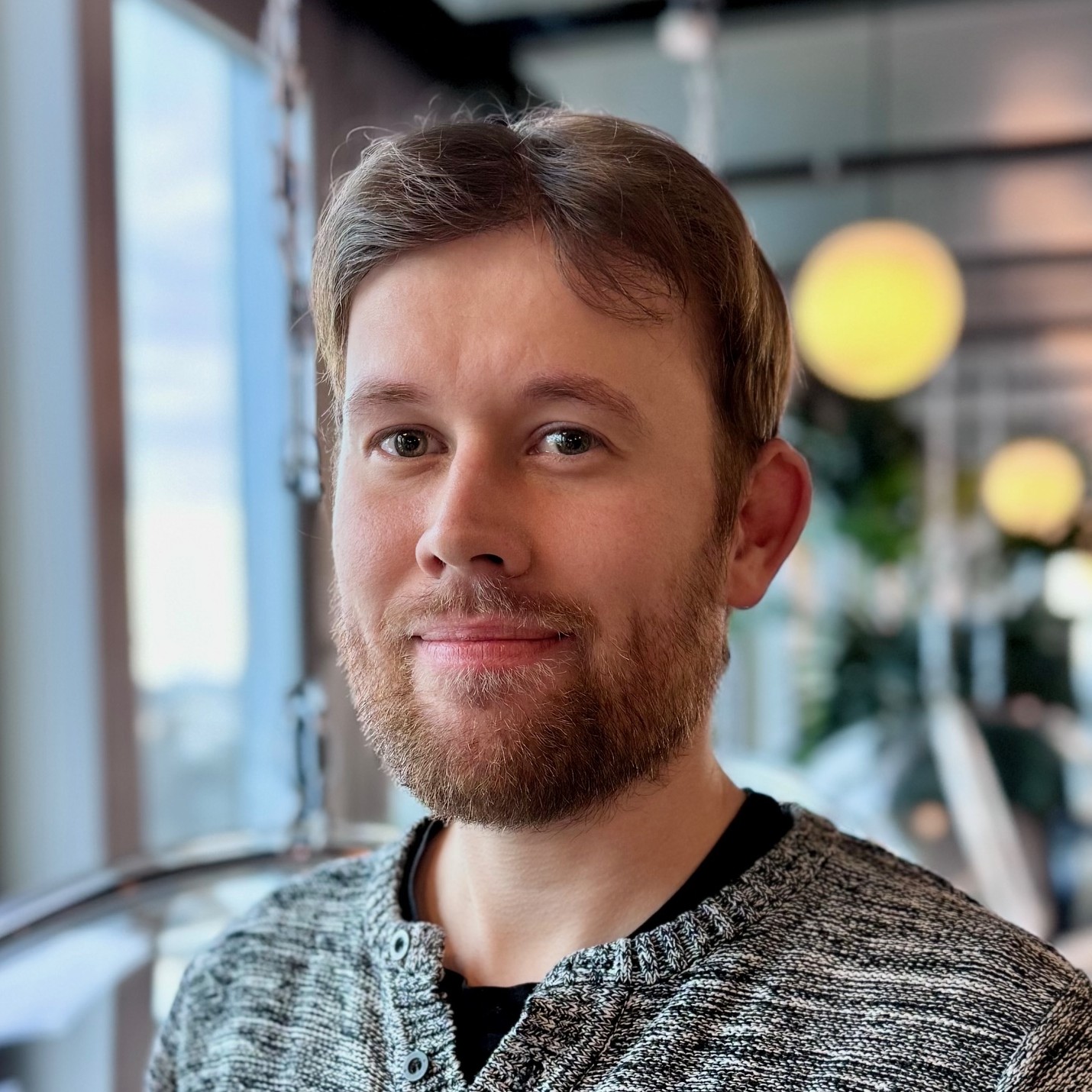 Philip trägt einen grau melierten Pullover, hat dunkelblonde Haare, trägt einen Vollbart und lächelt in die Kamera